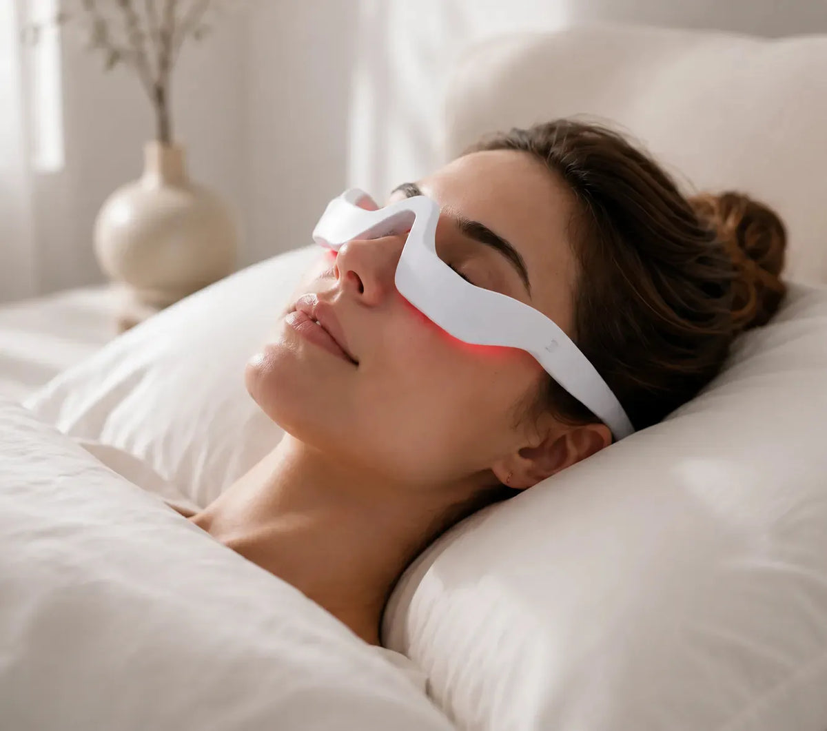 Woman relaxing on bed wearing white red light therapy eye glasses, at-home night skincare device to fade dark circles, reduce puffiness and fine lines, cozy minimalist bedroom background