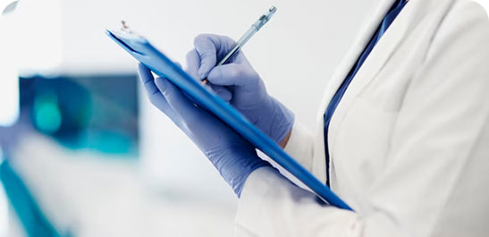 Person wearing a white coat and blue gloves holding a blue clipboard with a pen.
