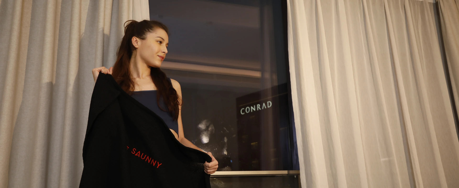 A woman with long hair stands near a window with beige curtains, holding a infrared sauna blanket insert with SUNNY logo on it.